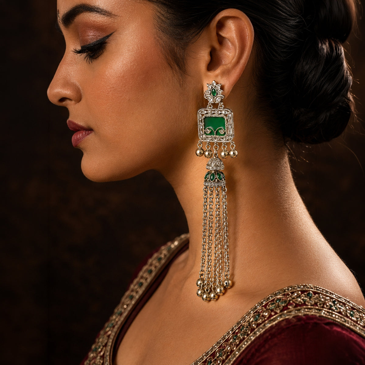 A woman in profile wearing a large traditional Indian jhumka earring with silver-toned metal, red enamel, and clear stones. The earring features a rectangular top with a red center, surrounded by clear stones and hanging dangling chains.