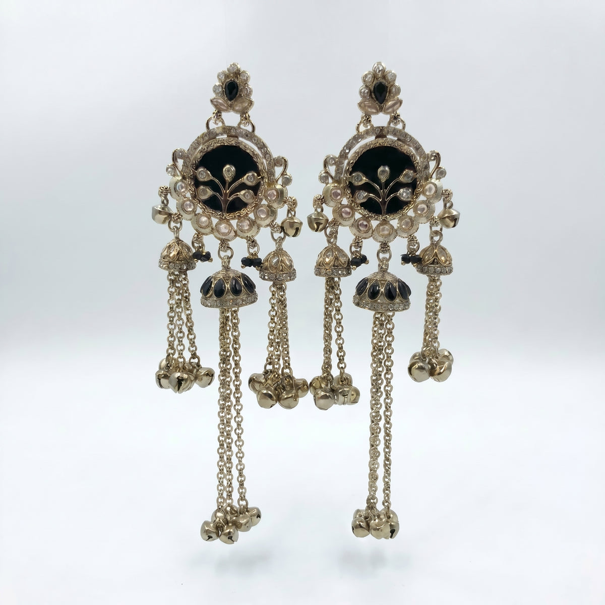 A pair of ornate Indian Kundan dangle earrings in gold-toned metal with black enamel, white faux pearls, and small clear stones, hanging against a light gray background.