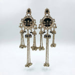 A pair of ornate Indian Kundan dangle earrings in gold-toned metal with black enamel, white faux pearls, and small clear stones, hanging against a light gray background.