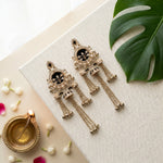 A pair of ornate gold-toned Indian-style earrings with black enamel details, white faux pearls, and dangling chain tassels, displayed on a textured cream-colored box with decorative props including a green monstera leaf, a brass diya, and small white flower buds.