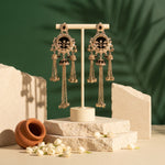 A pair of ornate gold-toned dangle earrings with black enamel and white stone detailing, displayed on a cream-colored T-stand. The stand is set on stacked stone blocks with a terracotta pot and white flowers in the foreground, against a dark green background with soft palm shadows.