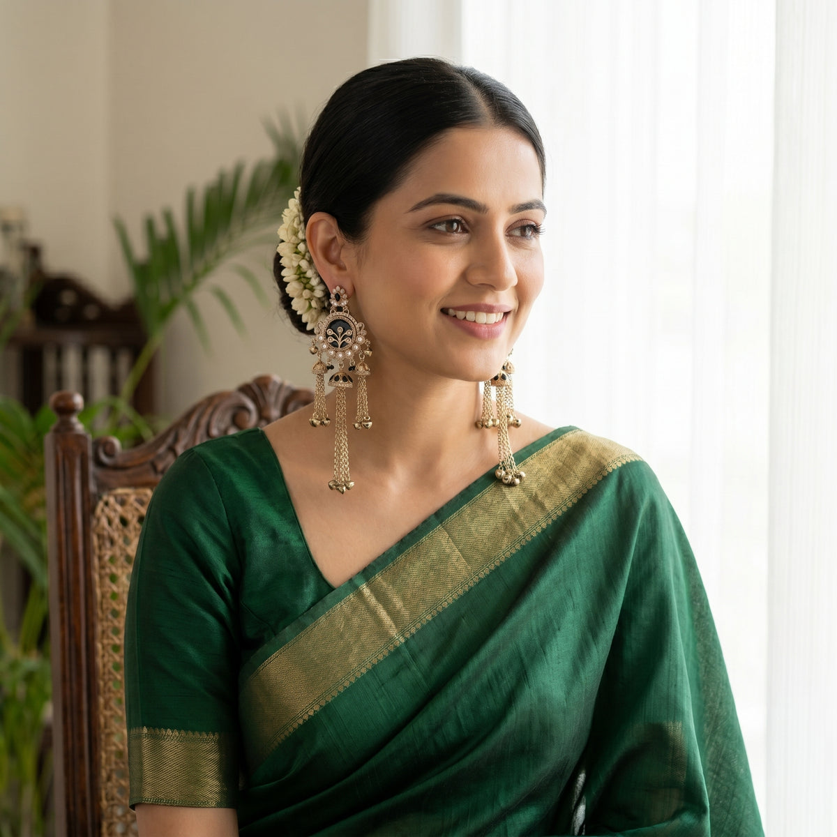 A woman wearing a dark green saree with a gold border, styled with traditional Indian jewelry and a hair ornament, seated in a wooden chair.