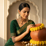 A woman in a dark green saree with gold borders is seen from the chest up, holding a terracotta clay pot decorated with yellow marigold flower garlands. She is wearing ornate gold earrings.