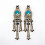 A pair of ornate silver-toned Indian jhumka earrings with turquoise blue enamel inlays, small pearl-like beads, and dangling bell chains, shown on a plain white background.