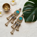 A pair of ornate gold-toned dangle earrings with turquoise blue stone centers and small blue accents, arranged on a light fabric surface with a lit diya, white flowers, and a large green leaf.