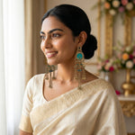 A woman wearing a cream-colored saree and matching blouse, showcasing a pair of ornate gold-toned dangle earrings with turquoise blue stones, in a softly lit indoor setting.