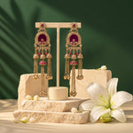 A pair of ornate gold-toned dangle earrings with deep red oval stones and small pink accents, displayed on a cream stand atop stone blocks with white lily flowers against a dark green background.