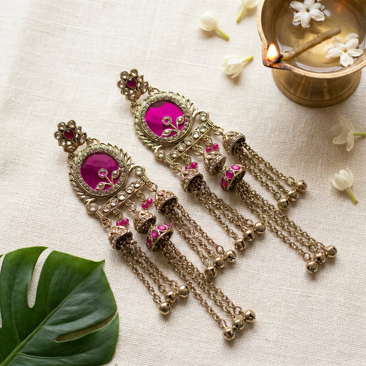 A pair of ornate Indian-style dangle earrings with bright pink stone centers, gold-toned filigree borders, and dangling bell charms, arranged on a light fabric surface with small white flowers, a green leaf, and a brass vessel in the background.