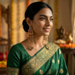 A woman wearing a dark green silk saree with gold zari embroidery and matching gold-toned traditional earrings, posed in a warm, decorated indoor setting.