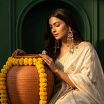 A woman in a cream-colored embroidered saree and ornate dangling earrings sits beside a large terracotta pot decorated with yellow marigold garlands against a dark green arched background.
