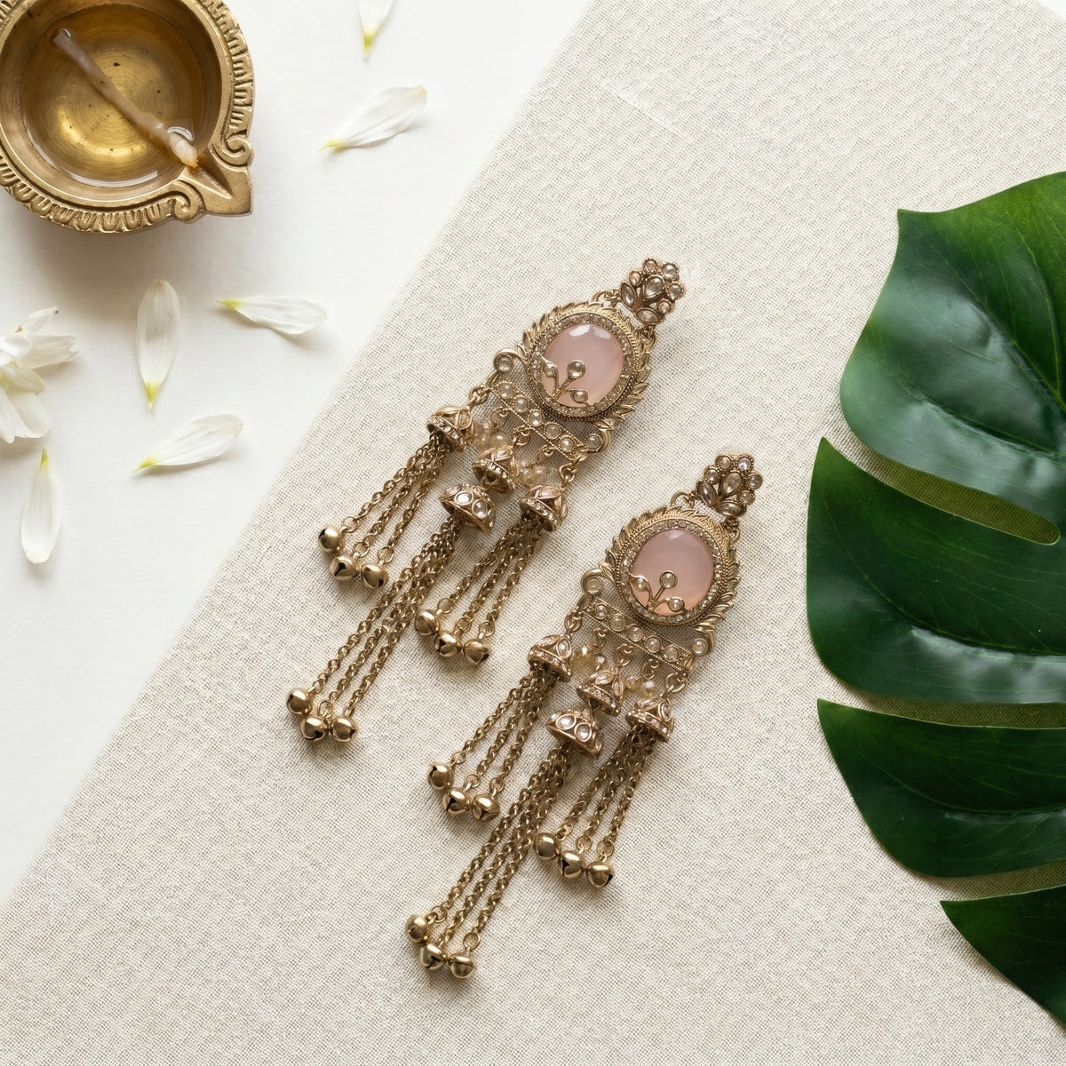 A pair of ornate gold-toned dangle earrings with round pale pink stone centers, arranged on a textured cream fabric alongside a brass diya, white flower petals, and a green monstera leaf.