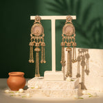 A pair of ornate gold-toned dangle earrings with oval pale pink stone centers and dangling chain bells, displayed on a white T-stand against a dark green backdrop with decorative props.