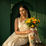 A woman in a cream-colored sari sits in a dark green room holding a ceramic vase filled with yellow and orange marigold flowers.