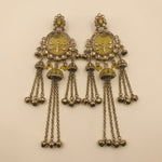 A pair of ornate silver-toned Indian chandelier earrings with yellow-green enamel inlays, floral motifs, and dangling bell charms, displayed on a plain beige background.