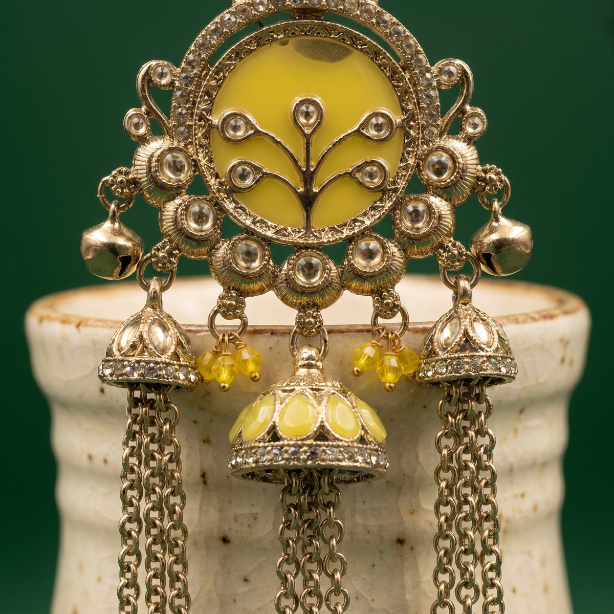 An ornate Indian-style gold-toned jhumka earring with a yellow enamel oval center, tree motif, and dangling bell-shaped charms with yellow enamel and bead drops, displayed on a ceramic stand against a dark green background.