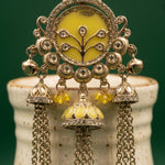 An ornate Indian-style gold-toned jhumka earring with a yellow enamel oval center, tree motif, and dangling bell-shaped charms with yellow enamel and bead drops, displayed on a ceramic stand against a dark green background.