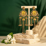 A pair of ornate gold-toned dangle earrings with yellow enamel panels and dangling chain bells, displayed on a cream jewelry stand. The stand is placed on stacked stone blocks with white flowers at the side, against a dark green leafy backdrop.