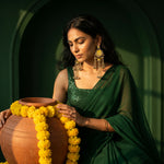A woman in a deep green saree and ornate gold-toned earrings holds a terracotta pot draped with yellow marigold flowers against a dark green arched background.