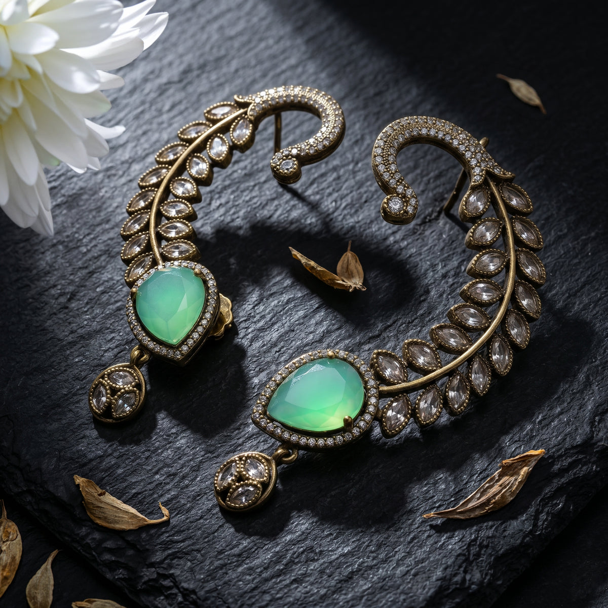 A pair of ornate gold-toned crescent-shaped earrings with large green teardrop gemstones and rows of clear stones, arranged on a dark textured surface with dried leaves and a white flower.