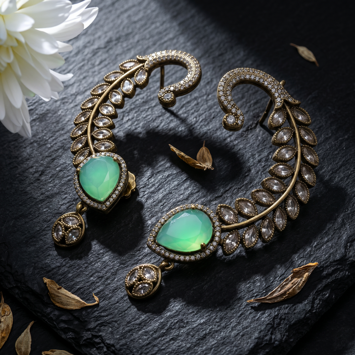 A pair of ornate gold-toned crescent-shaped earrings with large green teardrop gemstones and rows of clear stones, arranged on a dark textured surface with dried leaves and a white flower.
