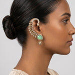 A close-up side profile of a woman wearing an ornate gold-toned ear cuff and matching drop earring with a light green teardrop stone and clear pavé details, against a plain light gray background.