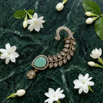 An ornate gold crescent-shaped brooch with a large oval light green stone and marquise-cut clear stones, styled on a dark green marble surface with white flowers.