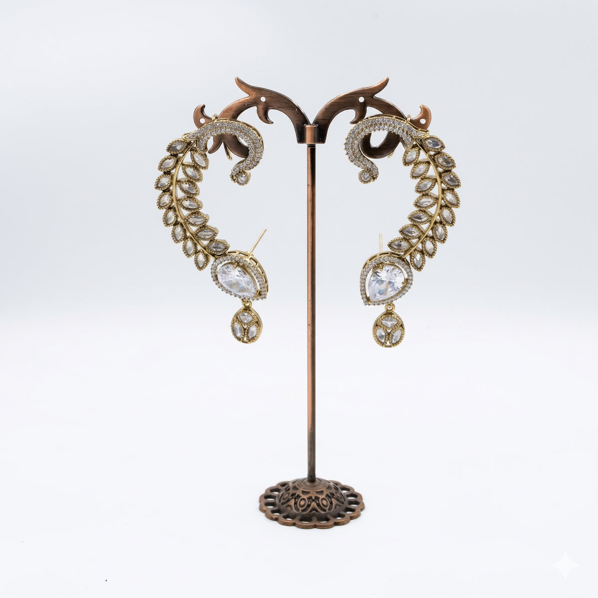A pair of ornate gold-toned drop earrings with clear rhinestone detailing and teardrop-shaped clear stones, displayed on a decorative bronze stand against a white background.