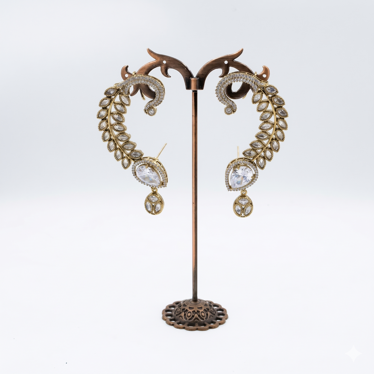 A pair of ornate gold-toned dangle earrings displayed on a decorative metal stand against a light gray studio background. Each earring features a leaf-like curved design set with clear rhinestones and a teardrop-shaped clear stone with a dangling clear stone below.
