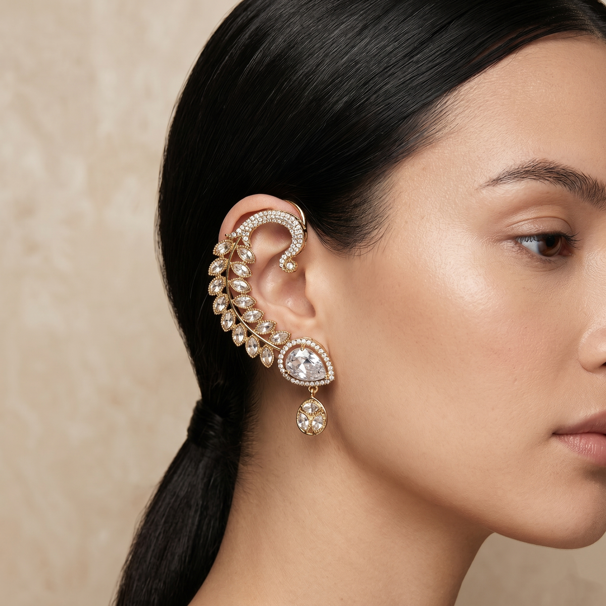 A close-up of a woman's ear wearing an ornate gold-toned ear cuff earring adorned with clear stones and a teardrop dangling charm, against a soft beige textured background.