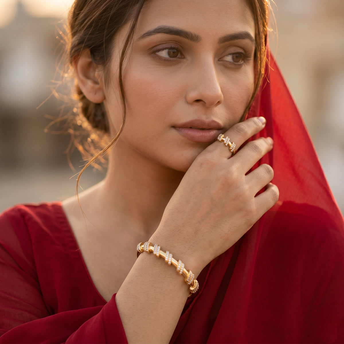 A woman in a red traditional outfit wearing a gold signet ring and a matching gold bracelet, captured in warm golden-hour lighting against a softly blurred background.