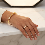 A close-up lifestyle shot of a person's hand resting on a white marble surface, wearing a gold T-bar bracelet and a matching gold ring with pavé-set clear stones.