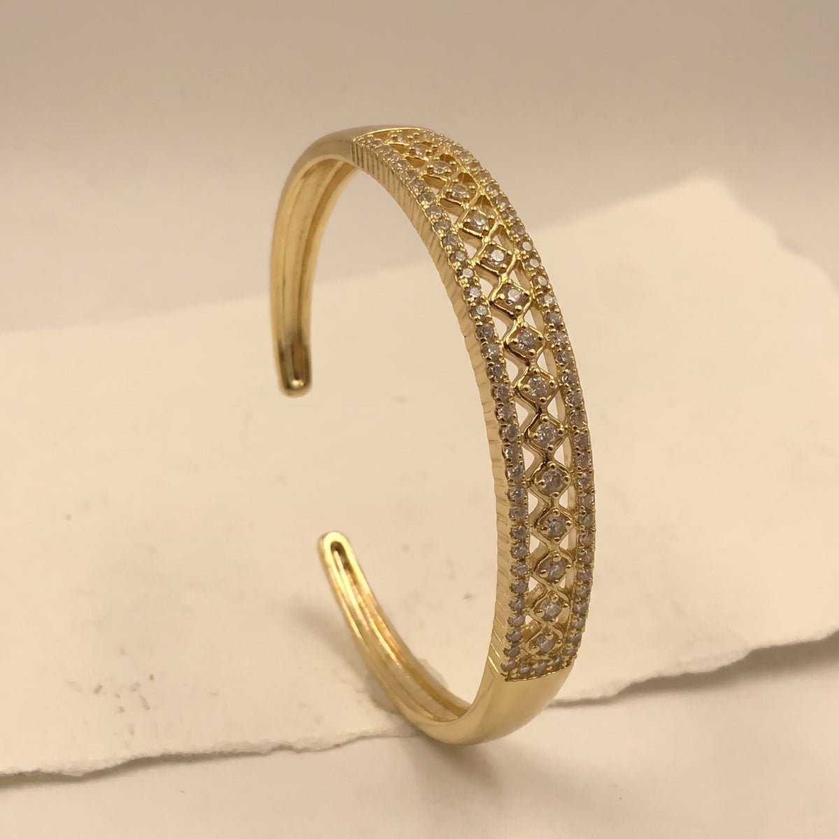 A gold-colored open cuff bangle bracelet featuring a detailed geometric pattern set with small clear stones, displayed on a textured off-white surface.