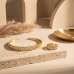 A gold cuff bracelet and matching ring with diamond-like pavé detailing, styled on a beige stone slab with warm neutral props.