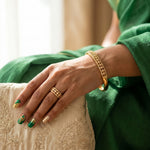 A close-up of a person's hand wearing a gold ring and a gold bangle, resting on a cream-colored embroidered cushion. The person is wearing a green traditional outfit, and the background is softly blurred.