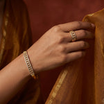A close-up of a person's hand wearing a gold bangle bracelet and matching ring, with the hand resting against a draped golden fabric against a warm reddish-brown background.