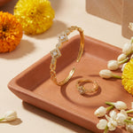 Gold jewelry set featuring a wide cuff bracelet and a matching ring, both adorned with clear pavé-set round stones, displayed on a terracotta tray with yellow and white flowers.