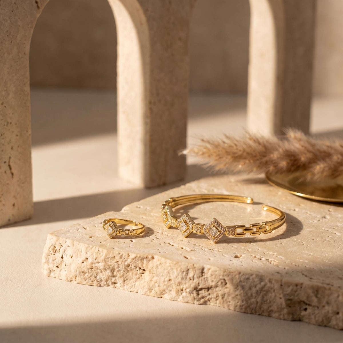 Gold jewelry set featuring a geometric link bracelet and matching ring, both adorned with small clear stones, displayed on a textured beige stone slab with warm architectural shadows.