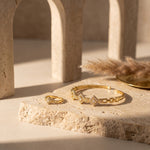 Gold jewelry set featuring a geometric link bracelet and matching ring, both adorned with small clear stones, displayed on a textured beige stone slab with warm architectural shadows.