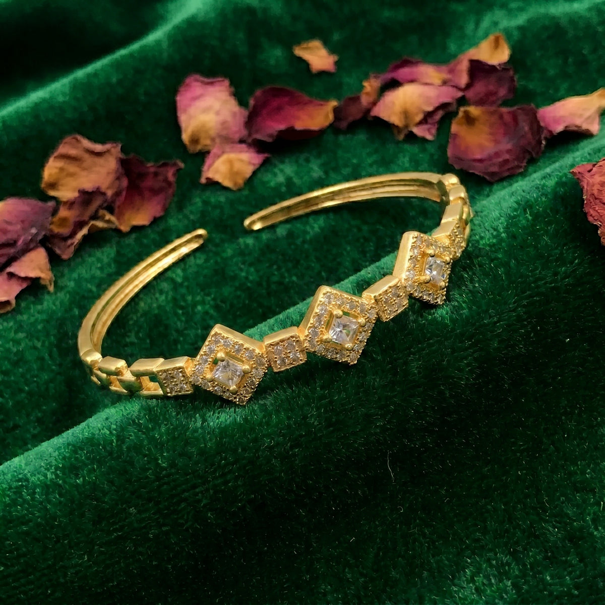 A gold-toned open cuff bracelet with four square diamond-shaped links, each set with small clear stones and a larger central clear stone, displayed on a dark green velvet fabric with scattered dried rose petals.