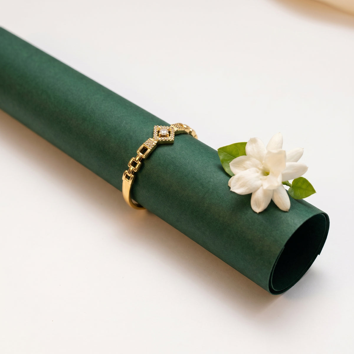 A gold chain bracelet with a diamond-patterned connector, displayed on a dark green paper roll alongside a white flower on a clean white background.