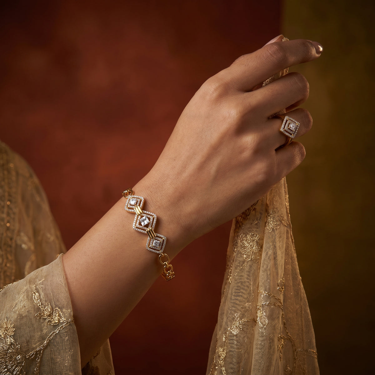A close-up of a person's hand wearing a gold bracelet with three linked square motifs, each set with a clear gemstone and a small gold heart at the center, paired with a matching ring. The person is wearing a beige embroidered garment against a warm, dark brown and gold background.