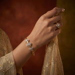A close-up of a person's hand wearing a gold bracelet with three linked square motifs, each set with a clear gemstone and a small gold heart at the center, paired with a matching ring. The person is wearing a beige embroidered garment against a warm, dark brown and gold background.