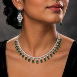 A close-up of a model wearing a silver-toned necklace and matching drop earrings set with emerald-cut green stones and clear stones, shown against a dark gray background.