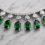 A close-up of a silver-toned tennis necklace with oval-cut emerald green gemstones and round clear stones, displayed on a white marble surface.