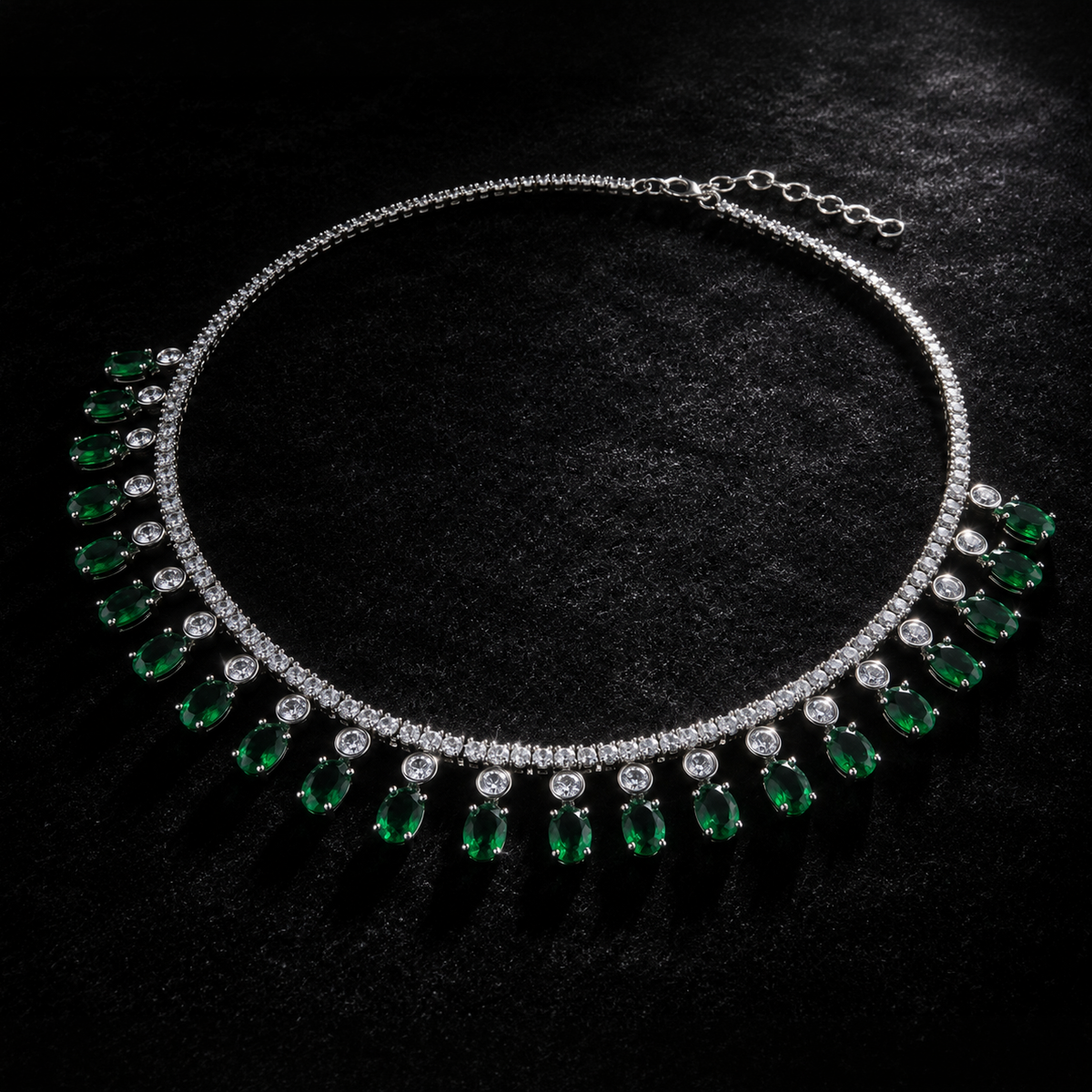 A silver-tone necklace featuring a row of green teardrop-shaped emerald-cut gemstones and clear round-cut stones, displayed on a textured black background.