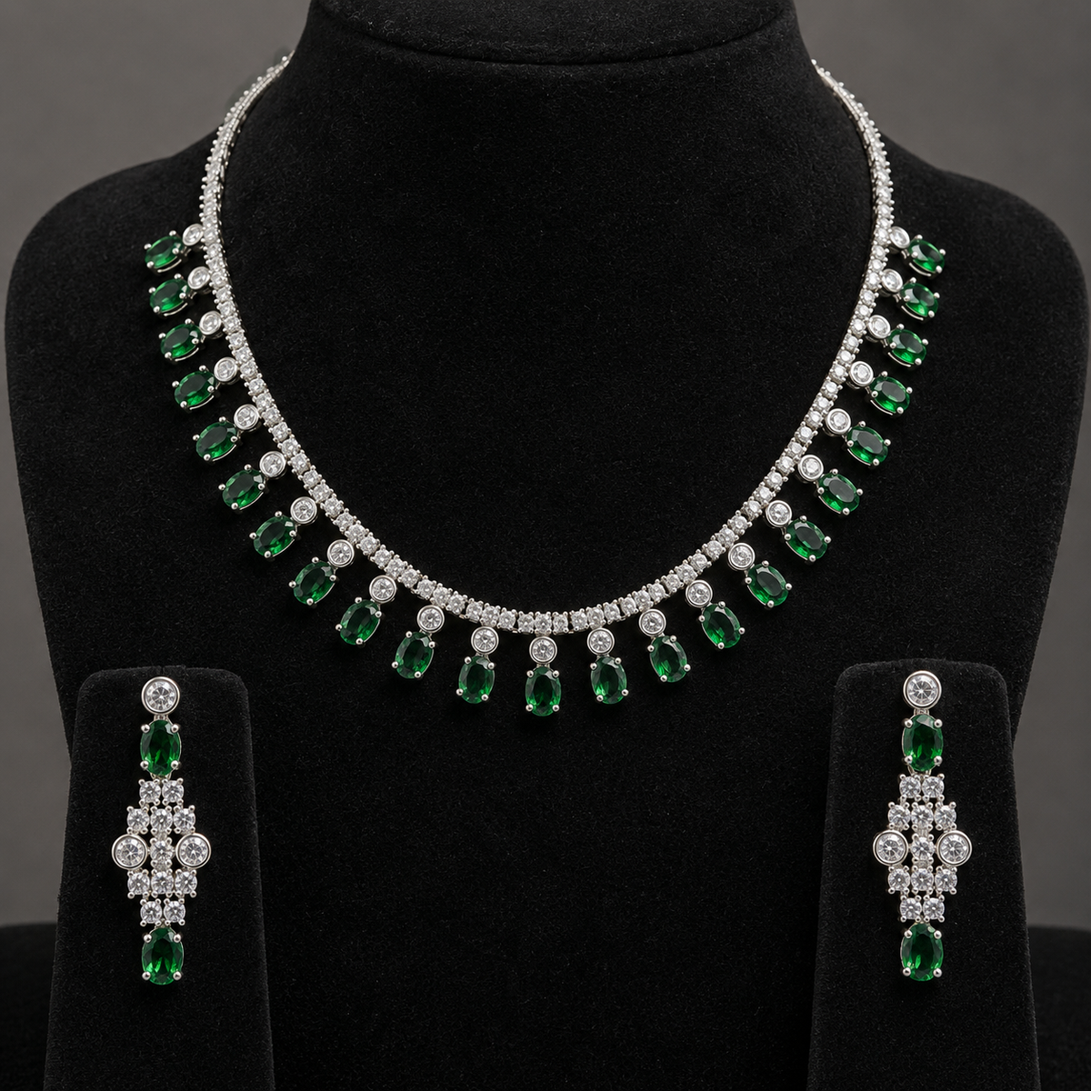A three-piece jewelry set featuring a silver-tone necklace and matching drop earrings with emerald-cut green gemstones and clear stones, displayed on a black velvet mannequin bust.