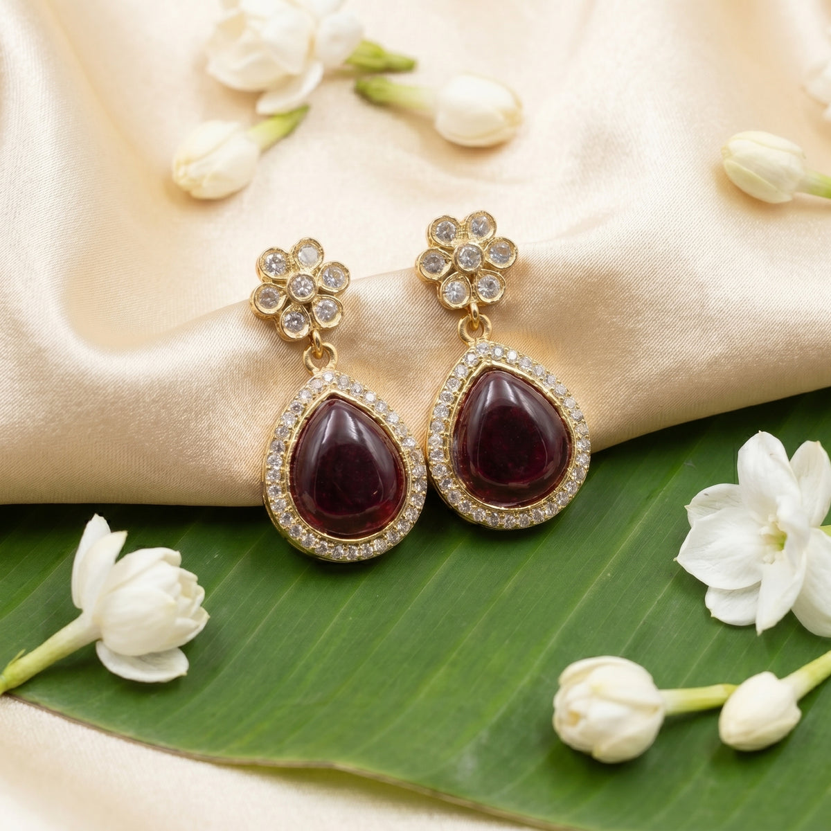 A pair of gold-plated dangle earrings with teardrop-shaped deep red gemstones surrounded by small clear stones, displayed on a beige satin fabric with white flowers and a green leaf.