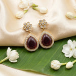 A pair of gold-plated dangle earrings with teardrop-shaped deep red gemstones surrounded by small clear stones, displayed on a beige satin fabric with white flowers and a green leaf.