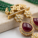 Gold-toned traditional Indian jewelry set featuring a decorative bar with floral motifs, a small green stone accent, and a teardrop-shaped drop with a deep red cabochon stone surrounded by clear crystals.