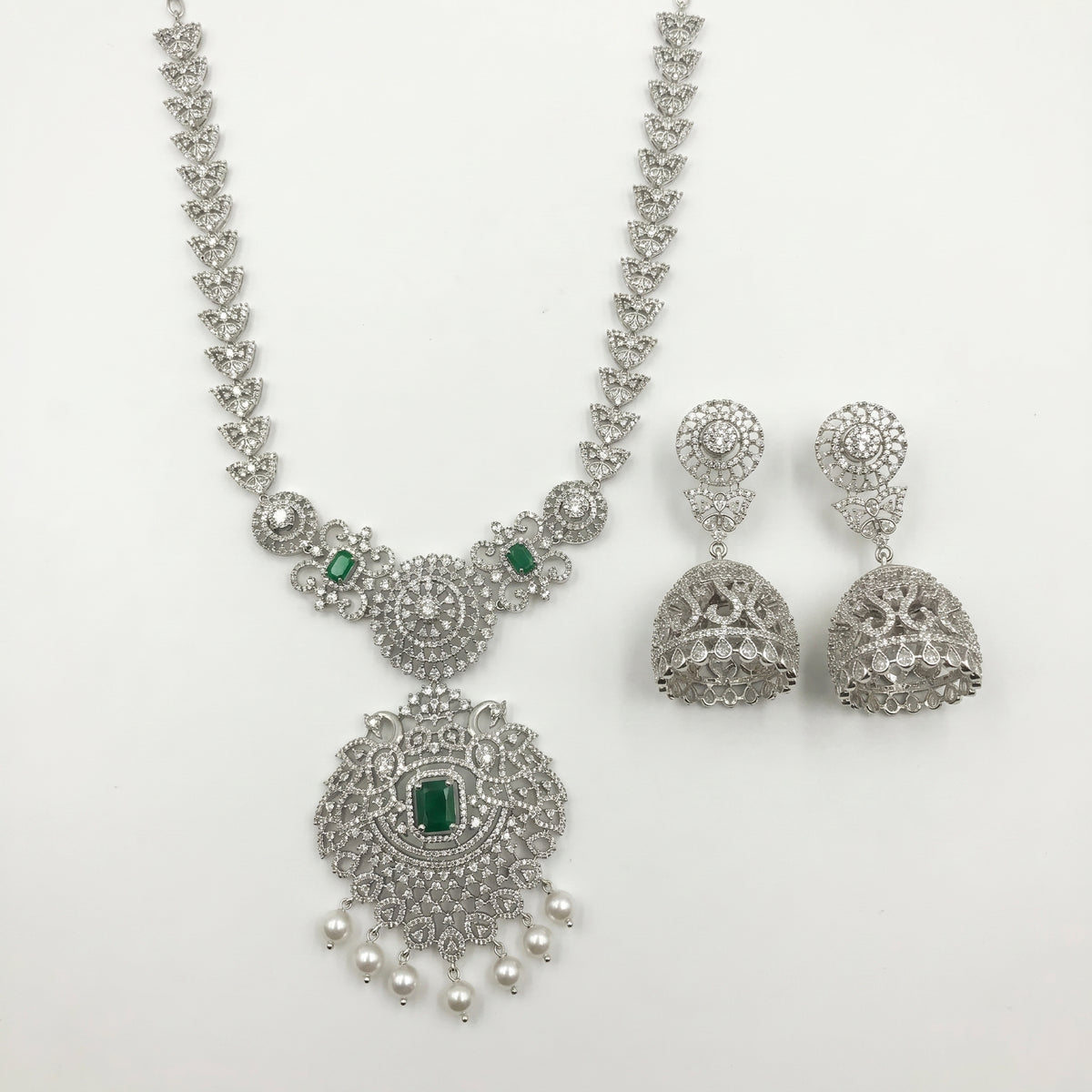 Silver-toned Indian jewelry set featuring a necklace and matching earrings, embellished with silver crystals, pearls, and small green emerald-cut stones, displayed on a clean white background.